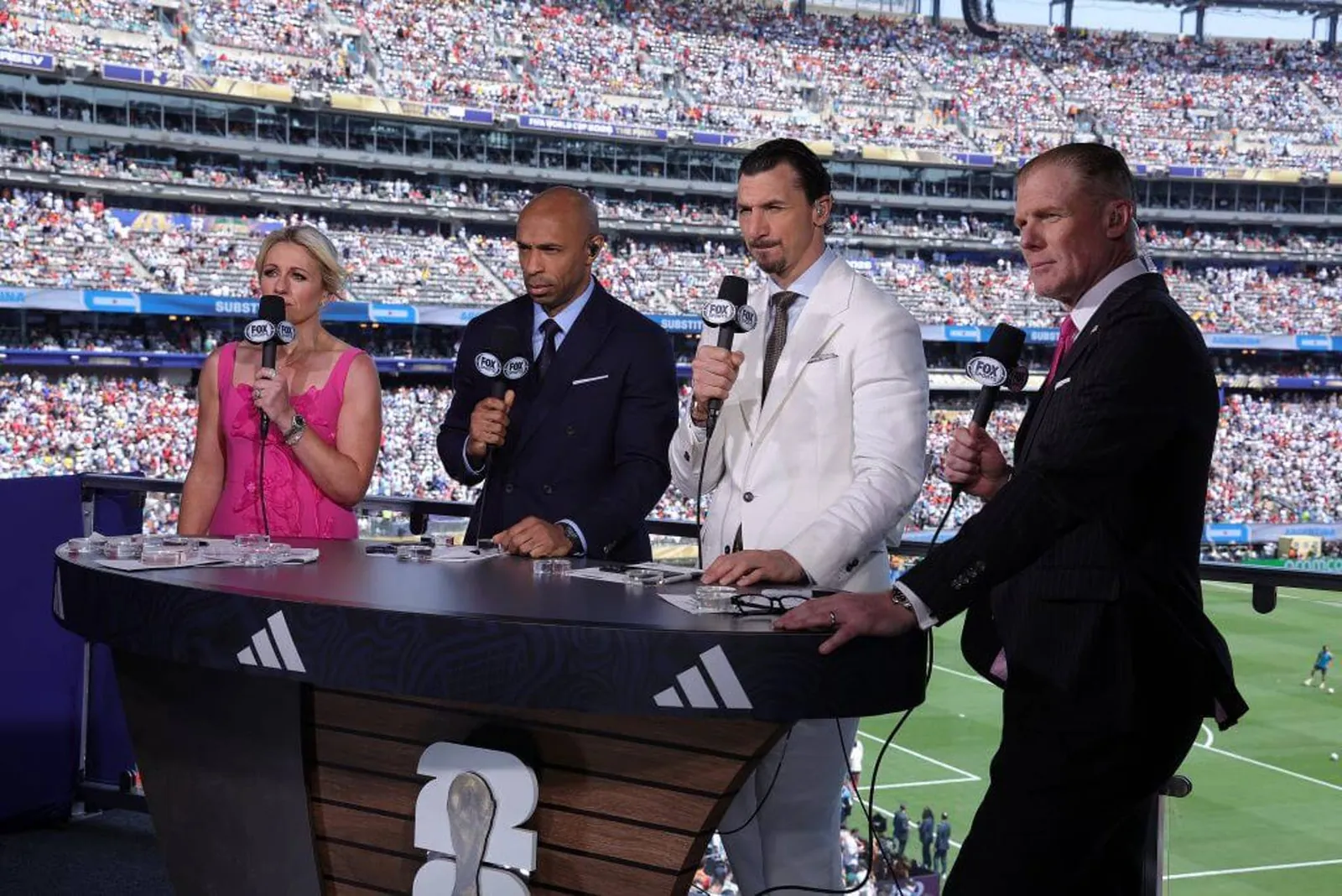 Rebecca Lowe, Thierry Henry, Zlatan Ibrahimović and Alexi Lalas stand at a desk with microphones with the stadium pitch and crowd in the background.